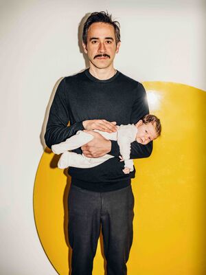 Een man kijkt met een glimlach de camera in, terwijl hij zijn baby in zijn armen houdt.