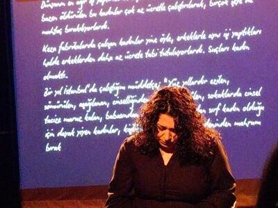 Elif Söylemez staat met gebogen hoofd op een podium. Achter haar worden handgeschreven Turkse teksten geprojecteerd op een paarsblauwe achtergrond.