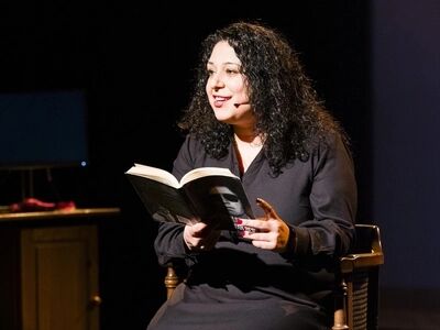 Elif Söylemez zit op een stoel op het podium en leest voor uit een boek. Ze draagt zwarte kleding en een headsetmicrofoon.