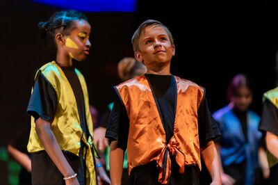 Twee kinderen in glanzende gekleurde vestjes staan op een podium; de jongen in het oranje vest kijkt trots omhoog, terwijl het kind naast hem naar links kijkt