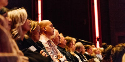 Publiek zit op de tribune van de Lindenbergzaal en luistert aandachtig naar een presentatie